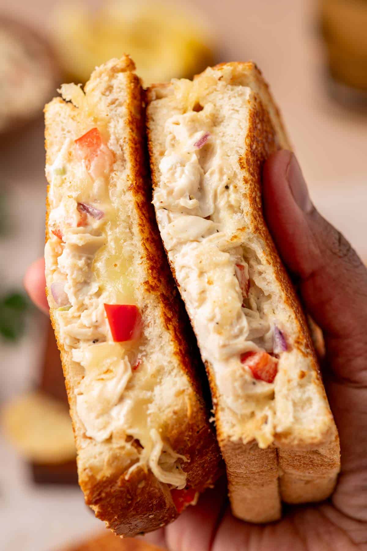 A perfectly grilled chicken salad melt sandwich a hand holding it , with cheese oozing from the sides and garnished with fresh parsley.