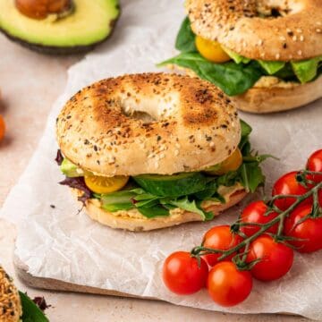 A vibrant veggie hummus bagel sandwich topped with creamy hummus, fresh avocado slices, crisp cucumber, juicy tomatoes, and mixed greens, served on a toasted everything bagel.