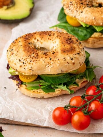 A vibrant veggie hummus bagel sandwich topped with creamy hummus, fresh avocado slices, crisp cucumber, juicy tomatoes, and mixed greens, served on a toasted everything bagel.