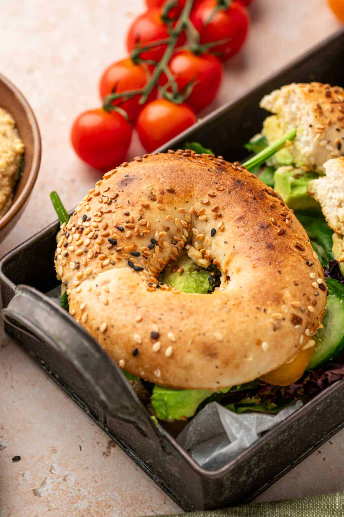 Creamy hummus spread on a toasted bagel topped with crisp veggies and avocado, served on a rustic tin tray.