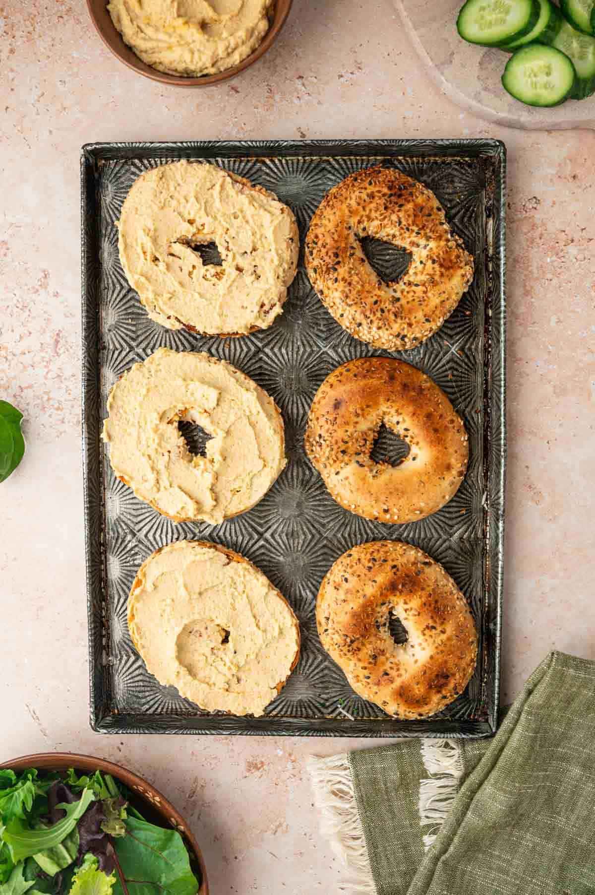 Hummus has been spreaded generously on the bagels on the baking tray.