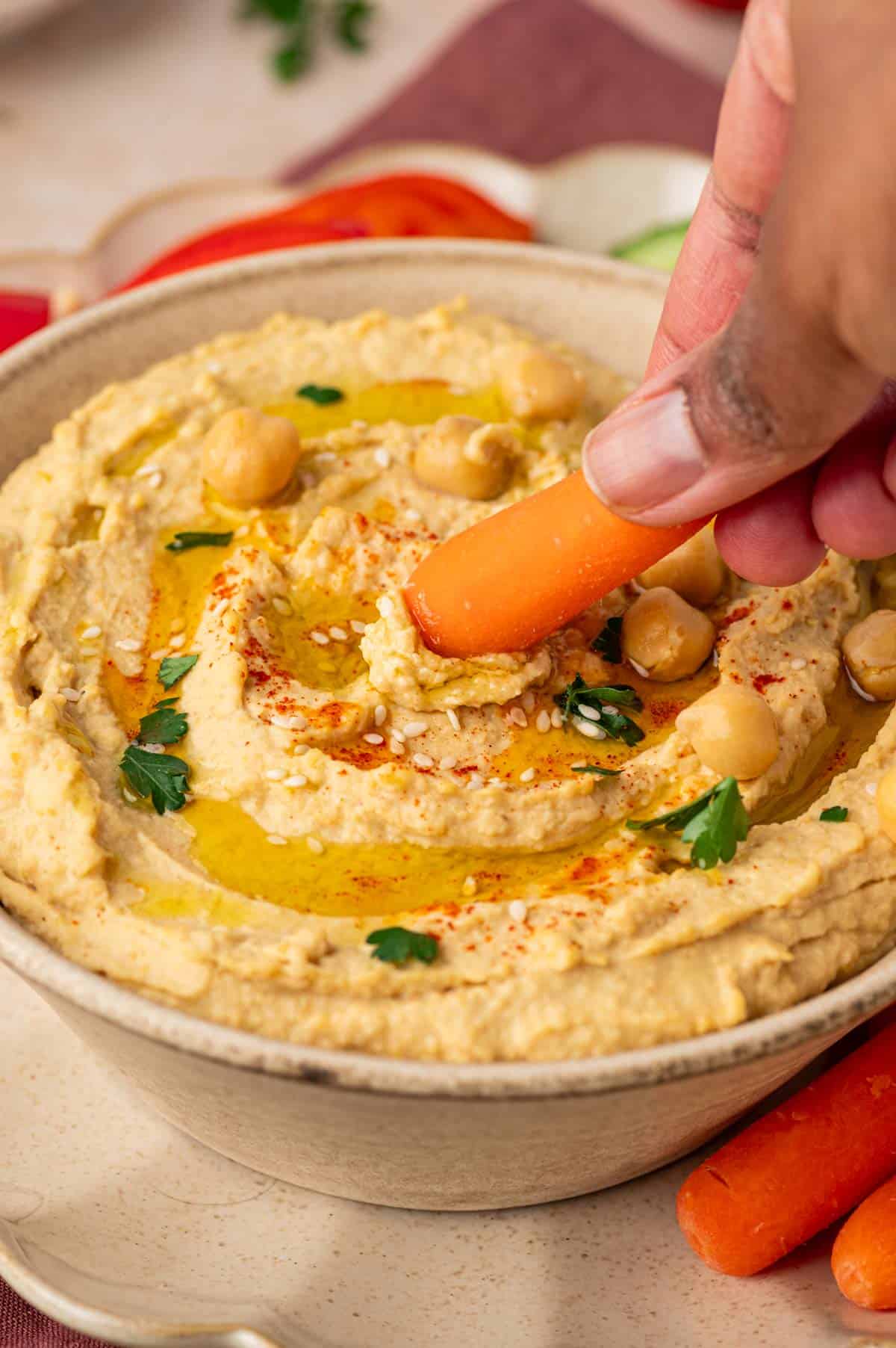 Garlic-free homemade hummus with a golden drizzle of olive oil, sprinkled with cumin and herbs, paired with crisp veggie dippers , a hand taking the hummus dip with a baby carrot.