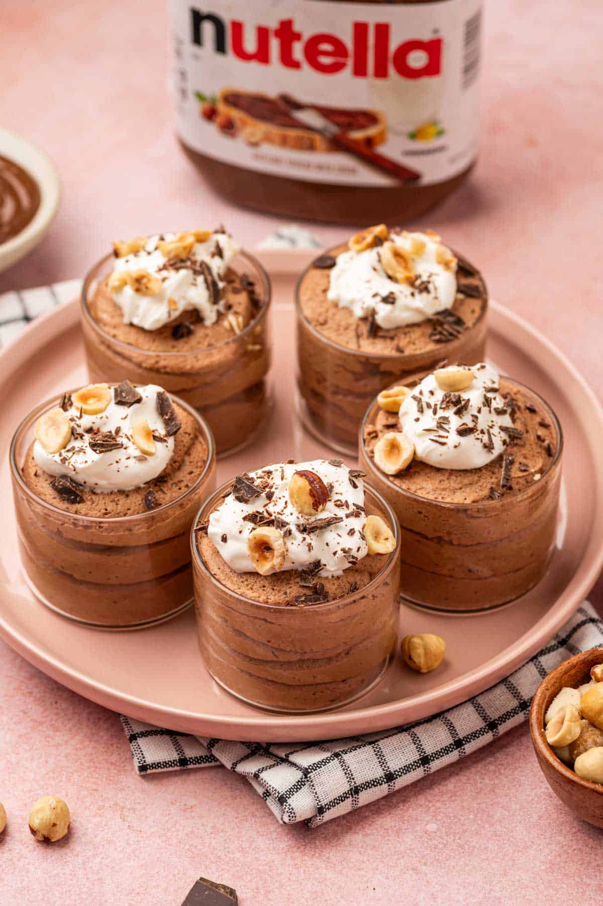 A dessert plate featuring Nutella mousse beautiful garnished with whipped cream and nuts. In the behind featuring Nutella jar on a pink background.