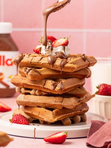 A stack of warm Nutella waffles served with a side of whipped cream, fresh strawberries, and a rich chocolate-hazelnut drizzle, creating a decadent and mouthwatering brunch treat.