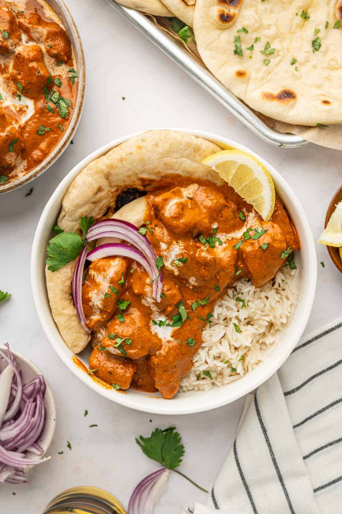 Top view of butter chicken in a bowl with naan and basmati rice. Garnished with cilantro and red onions.