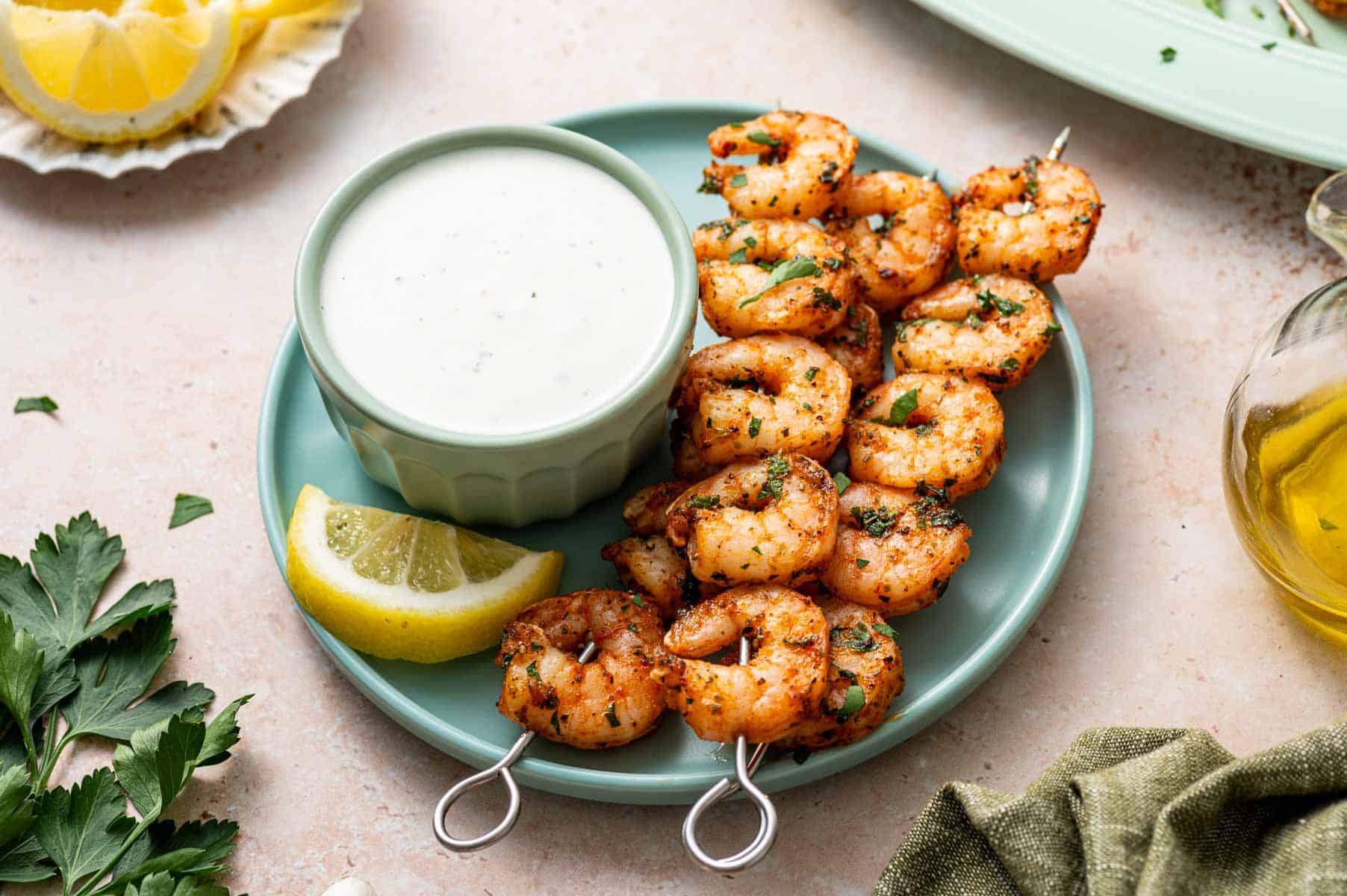 Close-up of oven-baked shrimp skewers arranged on a blue plate, garnished with fresh parsley and lemon wedges with ranch dip in a blue bowl.