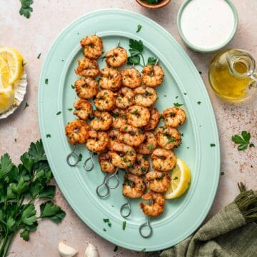 Golden roasted shrimp skewers fresh out of the oven, served with dipping sauce and herbs arranged beautifully on a green platter.