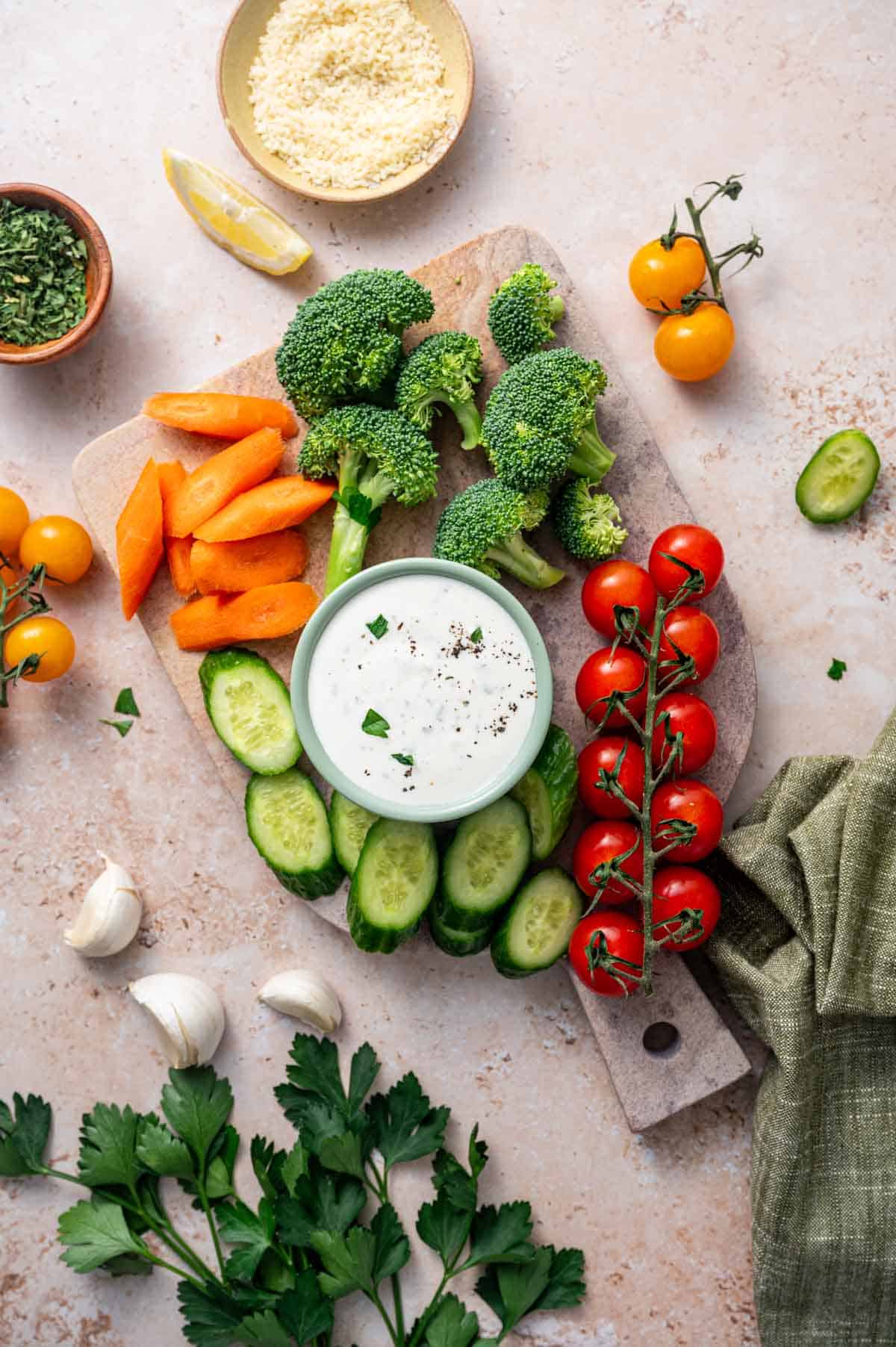 Overhead shot of Bowl of cheesy ranch dip made with buttermilk and fresh parmesan sorrounded by veggies like cucumber, carrots, broccoli, cherry tomatoes.
