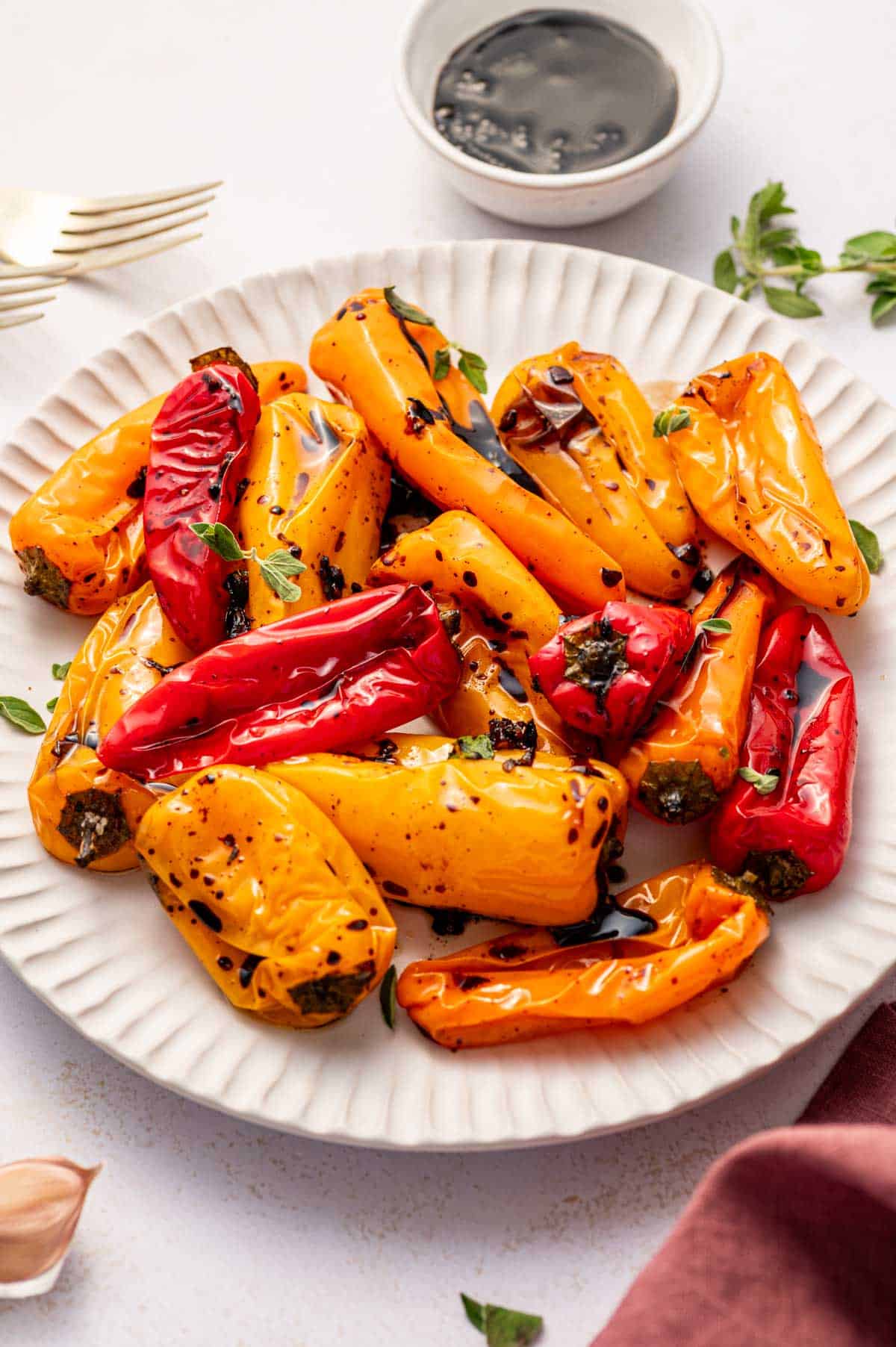 Colorful roasted red, yellow, and orange peppers served with fresh herbs in a white plate along with a small bowl of balsamic vinegar.