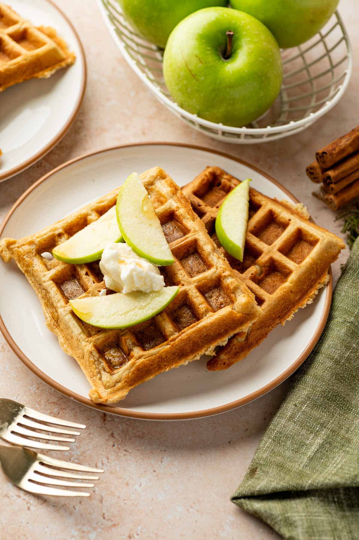 Golden brown cinnamon apple waffles on a white plate with fresh apple slices, whipped cream and maple syrup