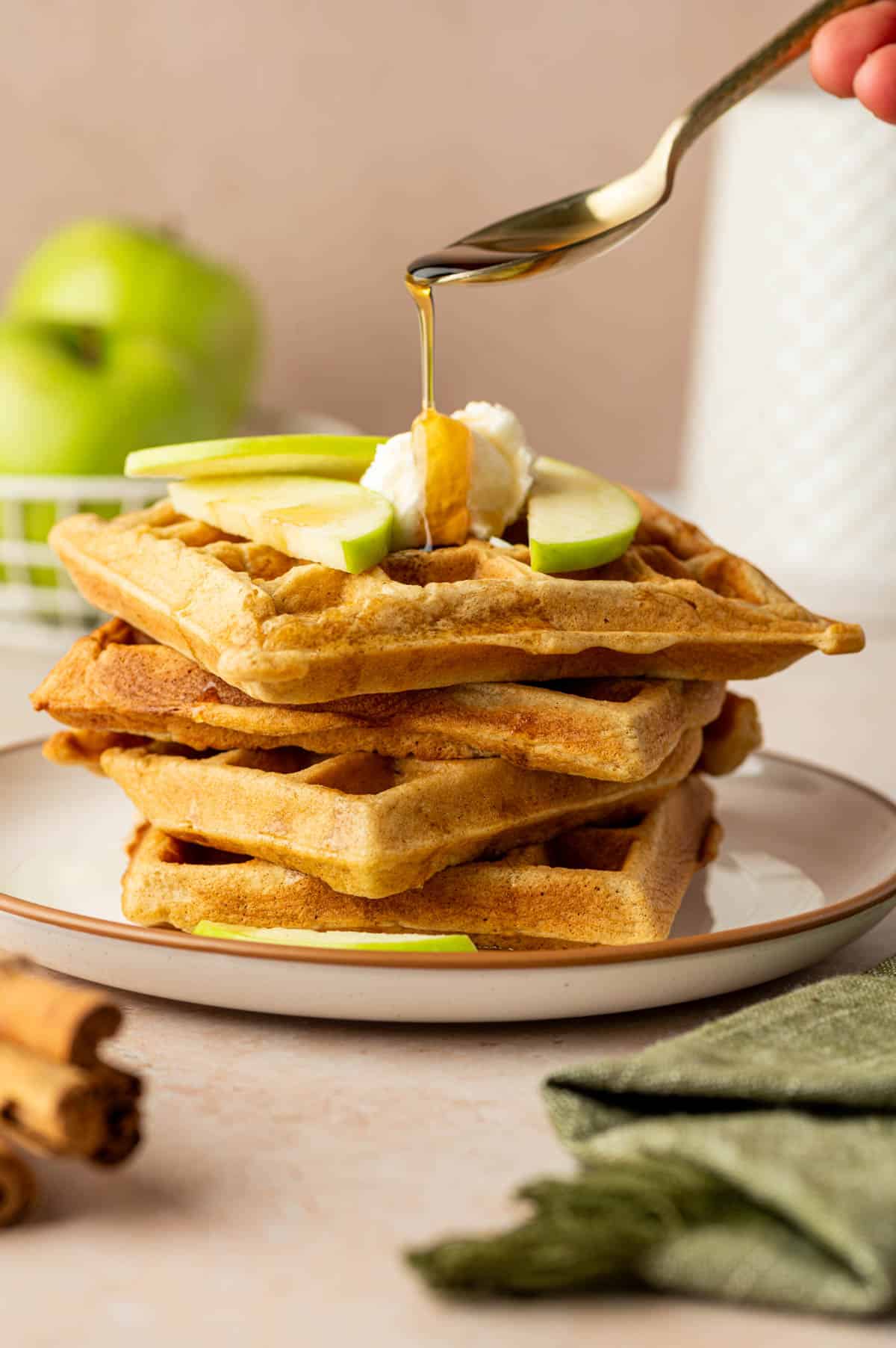 Stack of cinnamon apple waffles topped with fresh apple slices and maple syrup.