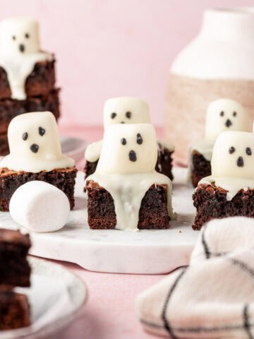 Several Halloween brownies topped with marshmallow ghosts, chocolate drizzle, and black eyes on a rustic marble tray.