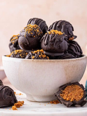 No-bake Biscoff truffles on a white bowl, coated in dark chocolate and drizzled with melted chocolate for a festive look. One truffle being cut opened.